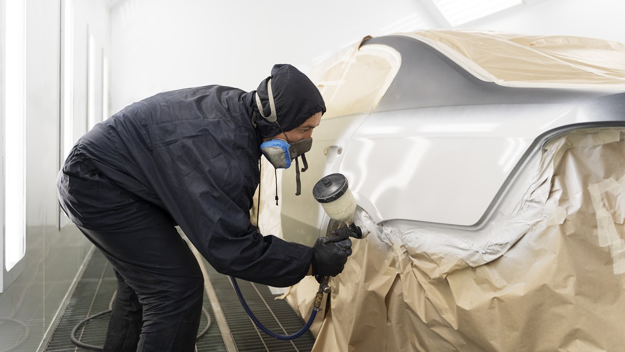 Seorang teknisi profesional menggunakan pakaian pelindung sedang melakukan proses cat full body mobil putih di dalam ruang oven bengkel.